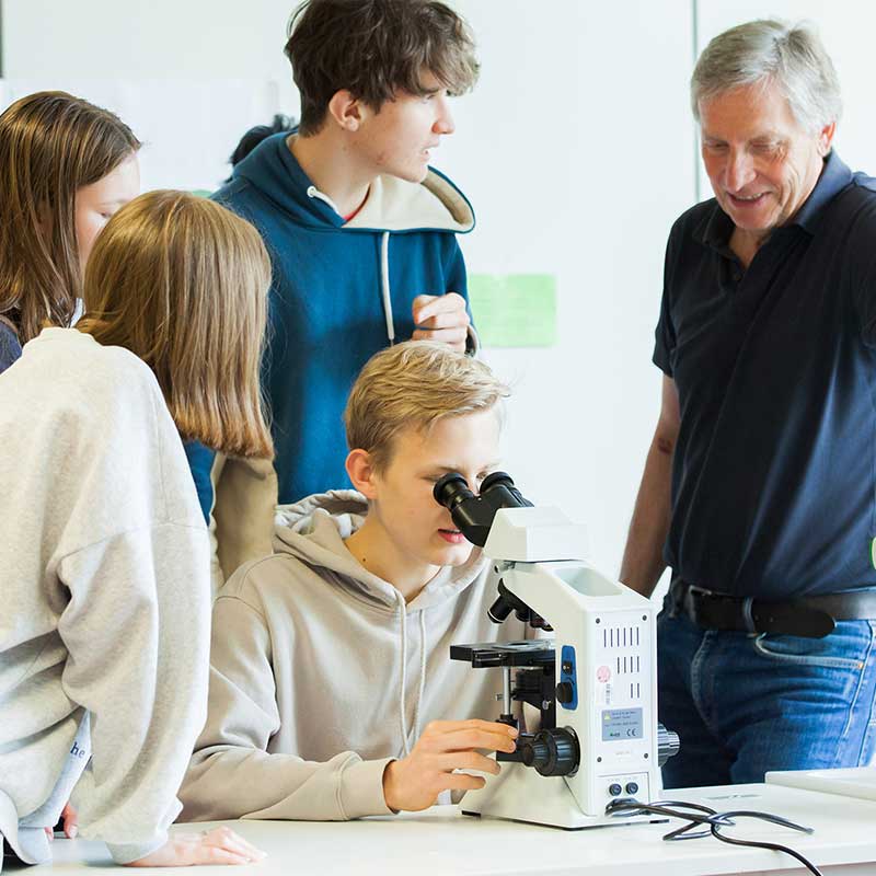 Schueler schaut durch Mikroskop, andere schauen ihm ueber die Schulter